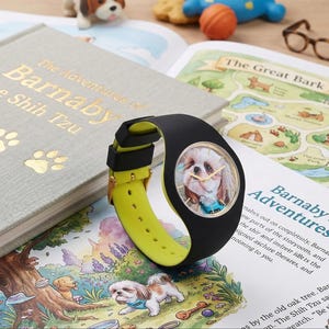 May include: A black and yellow wristwatch with a portrait of a Shih Tzu dog on the face. The watch is on top of a book titled "The Adventures of Barnaby the Shih Tzu". The watch band is yellow and black.