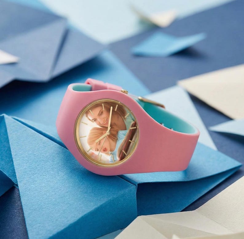 May include: A pink wristwatch with a gold-colored bezel and a light blue inner band. The watch face displays a photograph of two people. The watch is set against a backdrop of blue paper shapes.