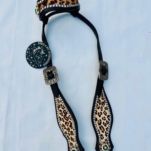 May include: A black and leopard print horse bridle with silver-tone buckles and decorative rhinestones. The bridle features a padded leopard print browband and matching cheek pieces, with a circular tag that reads "HORZE PET INTERNATIONAL".
