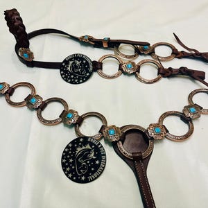 May include: Brown leather horse tack set with turquoise detailing and decorative metal rings. Includes a headstall, browband, and breast collar. Features a black circular tag with a horse graphic and the text "HORSE N PET INTERNATIONAL".