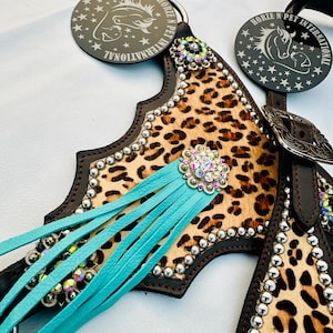 May include: A close-up of a horse tack set with a leopard print design. The set features brown leather, turquoise fringe, and silver studs. The tack includes decorative rhinestones and a metal buckle.