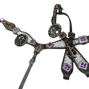 May include: A horse bridle with a black leather base, silver accents, and leopard print details. The bridle features purple square gemstones and a circular emblem with text and a horse head. The design includes buckles and straps.