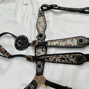 May include: A black and white patterned horse bridle with decorative studs and jewel accents. The bridle features a leather design with a metal buckle and a circular emblem with text and a horse graphic.