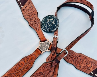 Puede incluir: Un juego de guarnicionería de cuero marrón para caballos con intrincados grabados florales y de cactus. El juego incluye un cabezal, un peto y riendas, todos adornados con detalles plateados y costuras decorativas. La guarnicionería presenta el logotipo "HORZE N PET INTERNATIONAL".