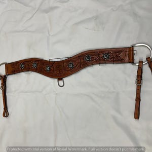 Könnte beinhalten: Ein brauner Leder-Brustgeschirr für Pferde mit floralen und scrollartigen Verzierungen. Der Kragen verfügt über silberfarbene Conchos und Beschläge, mit verstellbaren Riemen und einem D-Ring für Befestigungen. Das Leder hat eine mittelbraune Farbe.