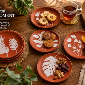 May include: A collection of terracotta-colored plates with white botanical designs. Some plates hold cookies, nuts, and fruit. The text "MADE FOR EVERY MOMENT" is visible. The plates are arranged on a wooden table, creating a warm, inviting aesthetic.