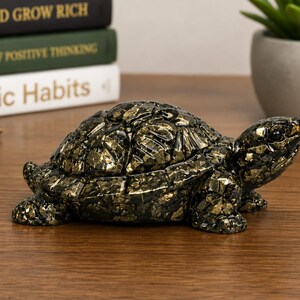 May include: A decorative turtle figurine with a mosaic-like pattern in black and gold. The figurine is on a wooden surface, with books and a pen in the background. The books have the titles "THINK AND GROW RICH", "THE POWER OF POSITIVE THINKING", and "Atomic Habits".