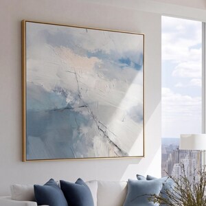 May include: A modern living room with a large abstract painting in a gold frame. A white sofa with blue pillows faces a glass coffee table. A striped armchair and a city view complete the scene.
