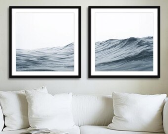 May include: Two framed prints featuring a minimalist depiction of ocean waves in shades of blue and gray. The artwork is set against a white background, framed in black, and displayed above a white sofa. The overall aesthetic is modern and serene.