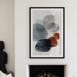May include: Abstract art print in a black frame. The artwork features overlapping shapes in shades of blue, gray, and rust against a light background. The print is displayed above a fireplace, with white furniture and decorative objects in the room.