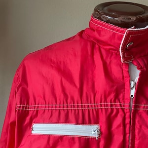 May include: A bright red jacket with a white zipper and pocket detail. The jacket has a stand-up collar with white trim and white stitching accents. The zipper is silver. The jacket is made of a shiny, lightweight material.