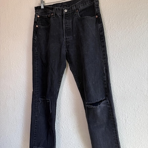 May include: Black denim jeans with distressed knee details and a raw hem. The jeans are hanging on a clear plastic hanger against a white wall. The jeans have a button fly and classic five-pocket styling.