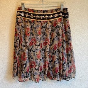 May include: A floral print skirt with a black waistband adorned with sequins and gold trim. The skirt features a pattern of red, blue, and tan flowers on a black background. The skirt is hanging on a clear hanger.