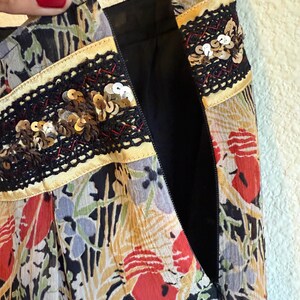 May include: Close-up of a floral print skirt with a black zipper. The skirt features a colorful pattern of red, blue, and yellow flowers on a black background. The waistband is decorated with black lace and gold sequins.