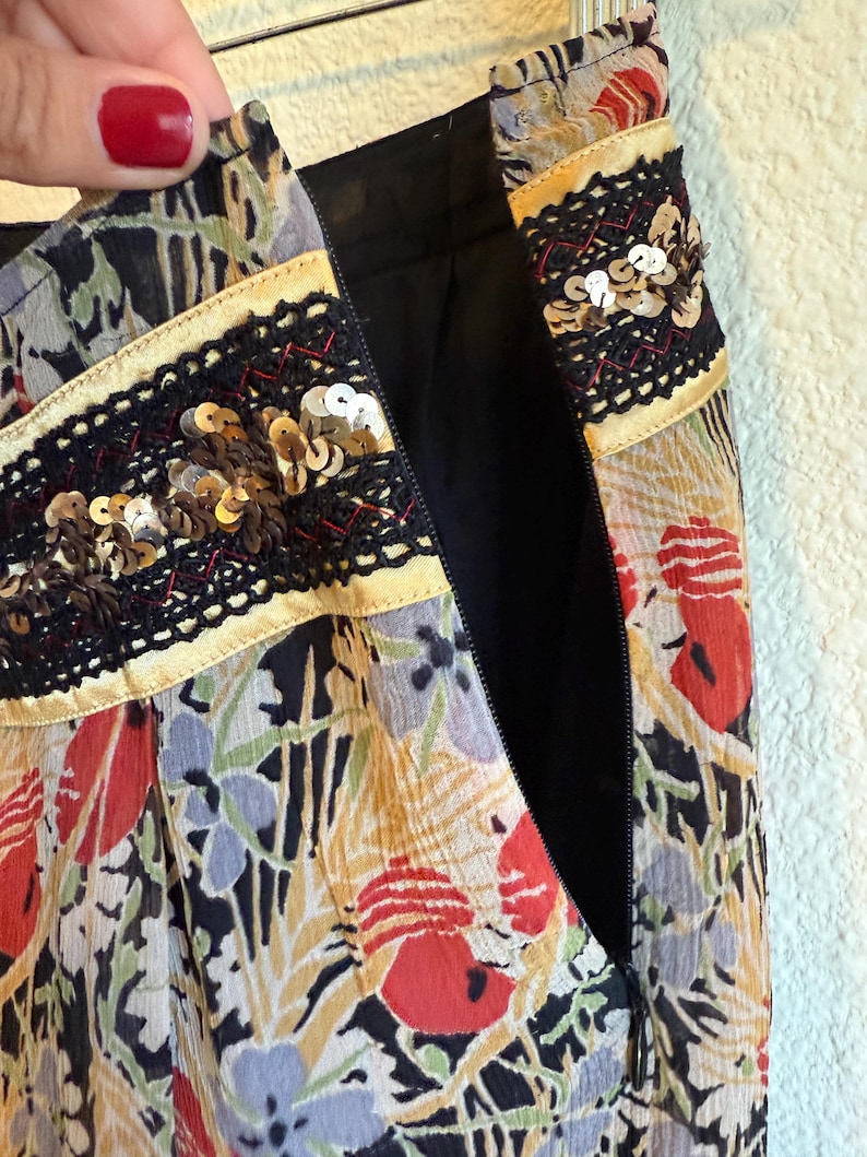 May include: Close-up of a floral print skirt with a black zipper. The skirt features a colorful pattern of red, blue, and yellow flowers on a black background. The waistband is decorated with black lace and gold sequins.