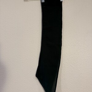 May include: A solid black scarf hanging on a clear plastic hanger against a white wall. The scarf is rectangular with a pointed end. The fabric appears to be a soft, flowing material.