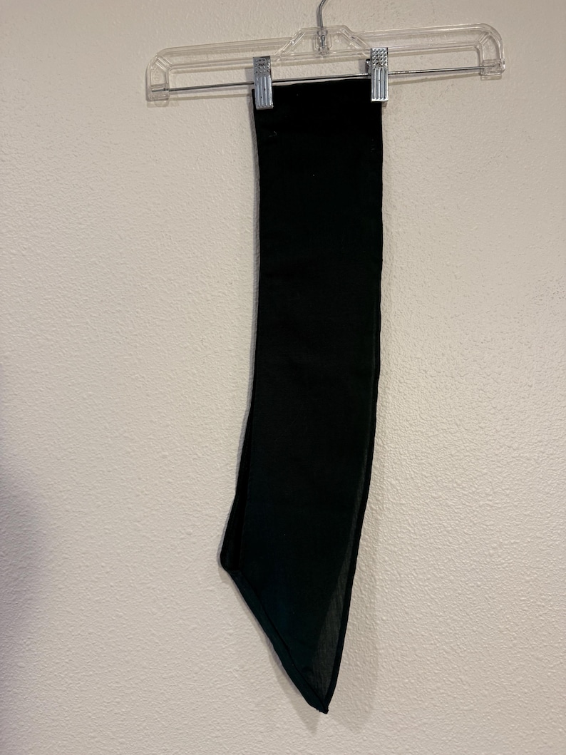 May include: A solid black scarf hanging on a clear plastic hanger against a white wall. The scarf is rectangular with a pointed end. The fabric appears to be a soft, flowing material.