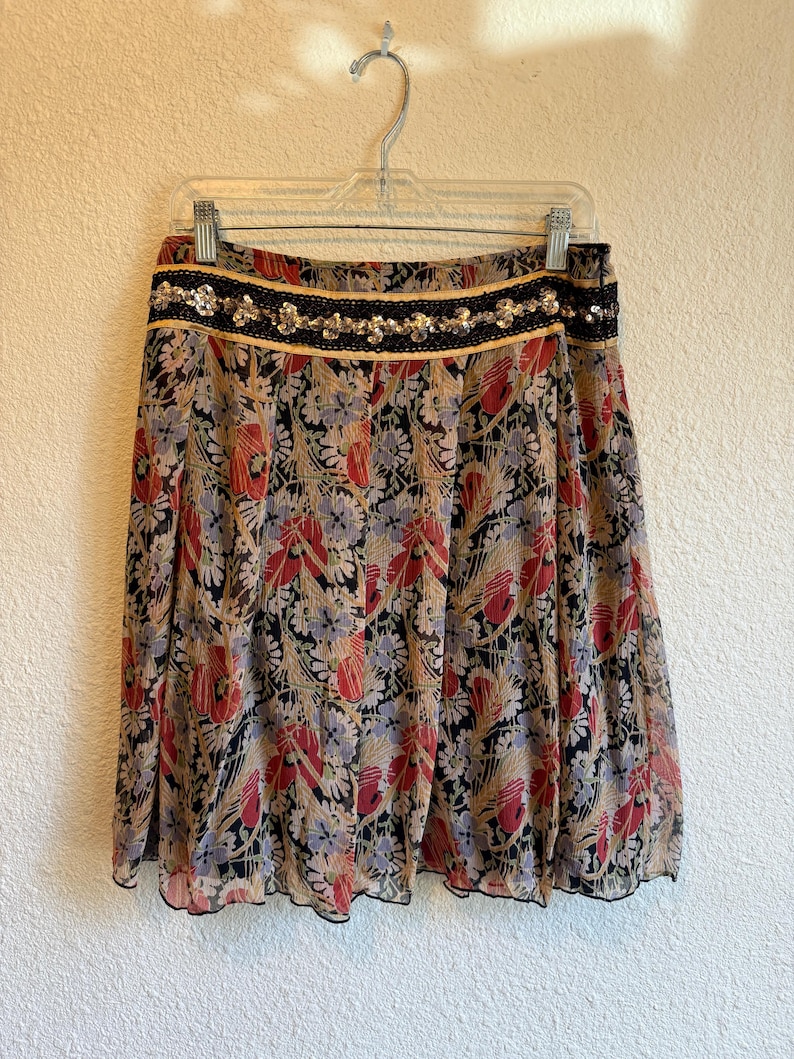 May include: A floral print skirt with a black waistband adorned with sequins and gold trim. The skirt features a pattern of red, blue, and tan flowers on a black background. The skirt is hanging on a clear hanger.