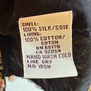 May include: A close-up of a fabric tag with the text "SHELL: 100% SILK/SOIE" and "LINING: 100% COTTON/COTON". The tag also includes washing instructions: "HAND WASH COLD, LINE DRY, NO IRON". The tag is beige with black text.