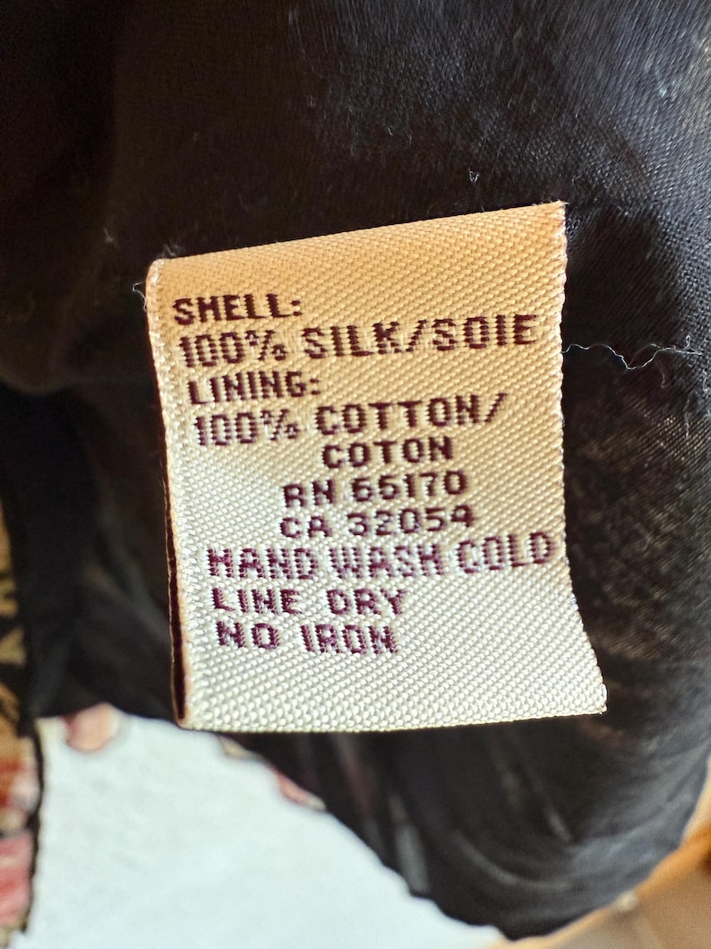 May include: A close-up of a fabric tag with the text "SHELL: 100% SILK/SOIE" and "LINING: 100% COTTON/COTON". The tag also includes washing instructions: "HAND WASH COLD, LINE DRY, NO IRON". The tag is beige with black text.