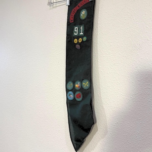 May include: A black sash with patches and the text "Western Rivers" and "91". The sash is hanging from a clear plastic hanger against a white wall. Patches include a star, a flower, and other colorful designs.