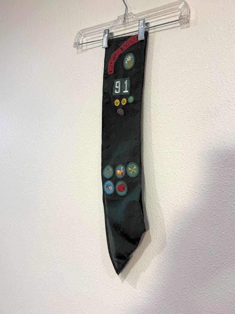 May include: A black sash with patches and the text "Western Rivers" and "91". The sash is hanging from a clear plastic hanger against a white wall. Patches include a star, a flower, and other colorful designs.
