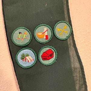 May include: Dark green fabric with five embroidered patches. The patches feature images of running figures, cooking utensils, crossed keys, a tent, and a red box. The patches are round with green borders.