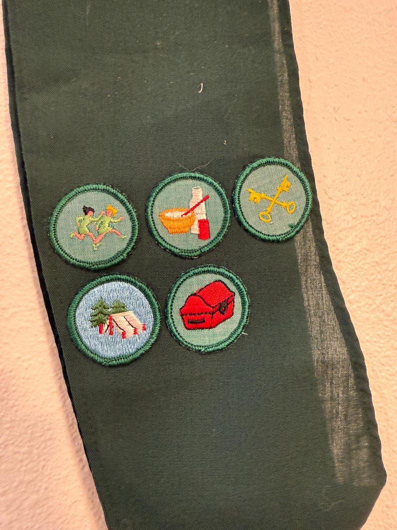 May include: Dark green fabric with five embroidered patches. The patches feature images of running figures, cooking utensils, crossed keys, a tent, and a red box. The patches are round with green borders.