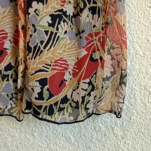 May include: Close-up of a sheer fabric with a floral pattern. The design features red, black, yellow, and green botanical elements on a beige background. The fabric has a slightly crinkled texture and a black trim.
