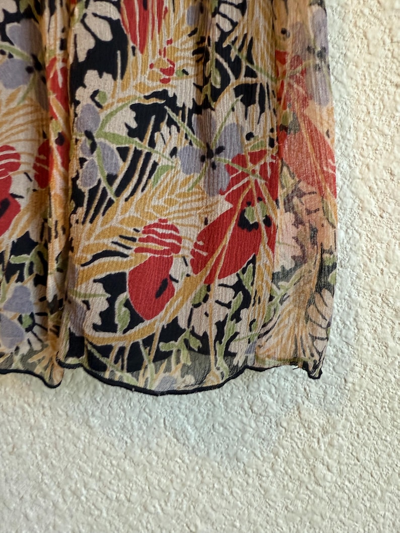 May include: Close-up of a sheer fabric with a floral pattern. The design features red, black, yellow, and green botanical elements on a beige background. The fabric has a slightly crinkled texture and a black trim.