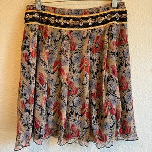 May include: A floral print skirt with a black, red, and beige pattern. The skirt has a black waistband with gold trim and sequin embellishments. The skirt is hanging on a hanger.