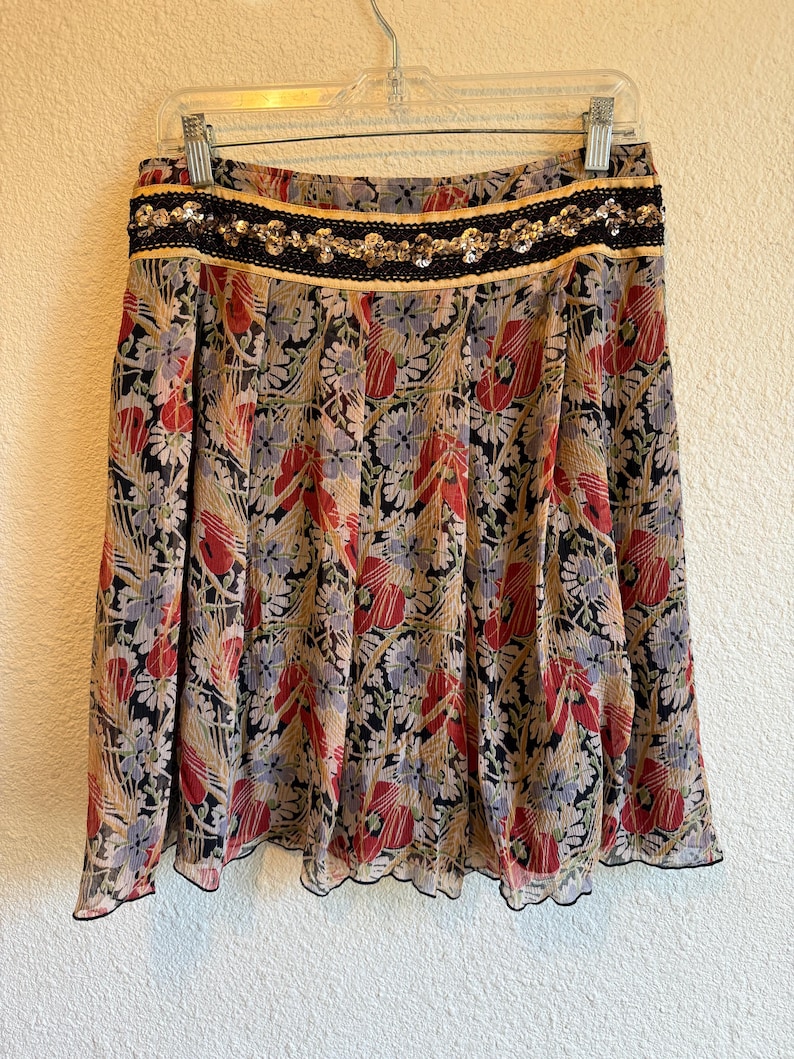 May include: A floral print skirt with a black, red, and beige pattern. The skirt has a black waistband with gold trim and sequin embellishments. The skirt is hanging on a hanger.