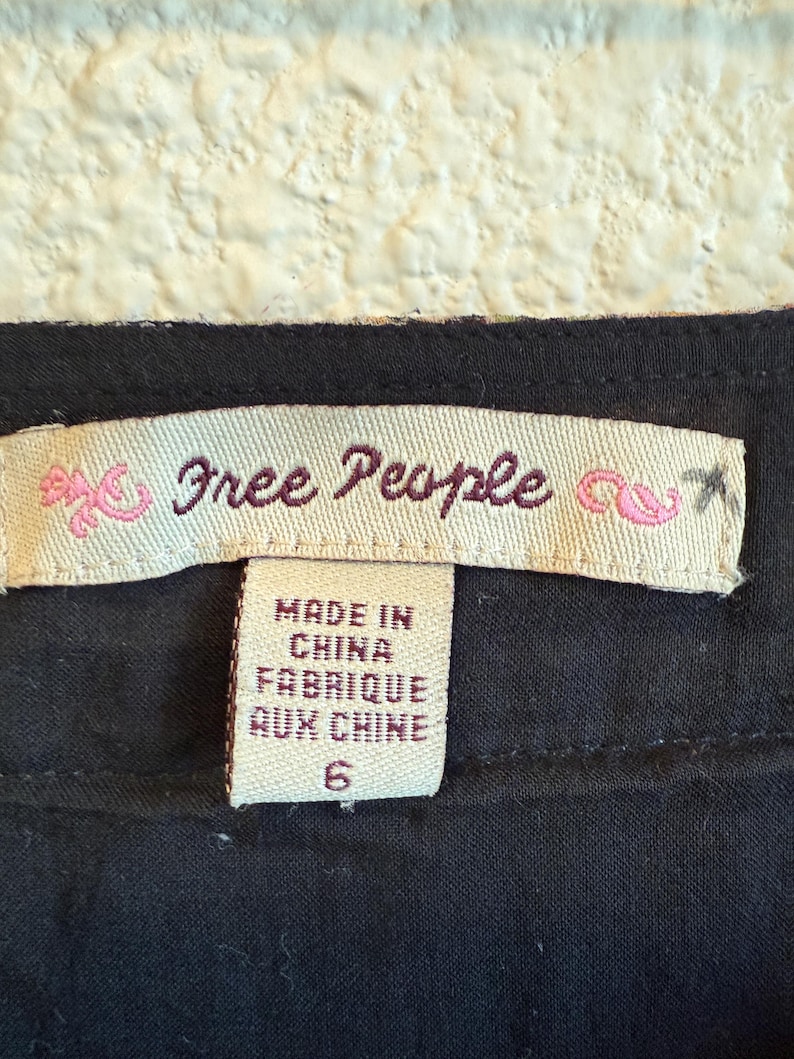 May include: Close-up of a clothing label with the brand name "Free People" in cursive script. The label also states "Made in China Fabrique aux Chine" and the size "6". The label is on a black fabric.