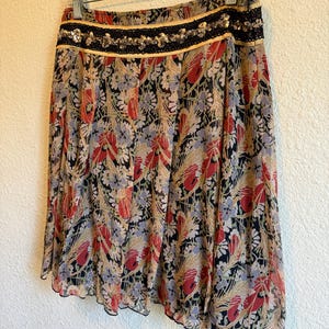 May include: A floral print skirt with a black, red, and beige pattern. The skirt has a black lace and sequin waistband and a scalloped hem. The skirt is hanging on a clear hanger against a white wall.