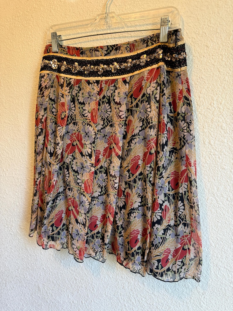 May include: A floral print skirt with a black, red, and beige pattern. The skirt has a black lace and sequin waistband and a scalloped hem. The skirt is hanging on a clear hanger against a white wall.