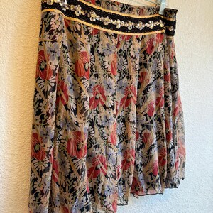 May include: A flowing skirt with a floral pattern in red, black, and beige. The skirt has a black sequined waistband with gold trim and is hanging on a white hanger. The fabric appears to be lightweight and semi-sheer.