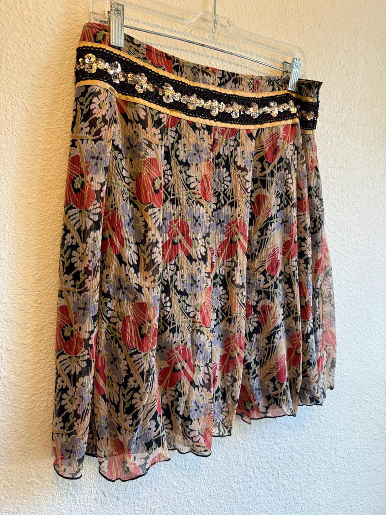 May include: A flowing skirt with a floral pattern in red, black, and beige. The skirt has a black sequined waistband with gold trim and is hanging on a white hanger. The fabric appears to be lightweight and semi-sheer.