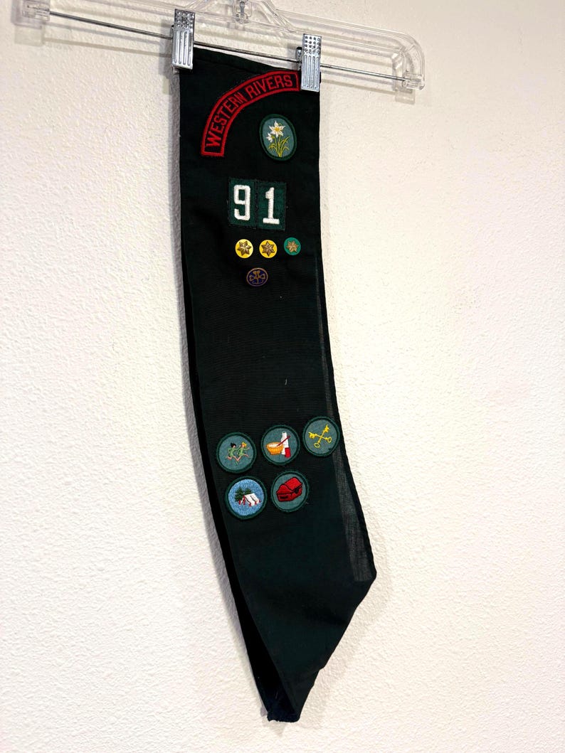May include: A dark green sash with patches and the text "WESTERN RIVERS" and "91". The sash has various patches with images of flowers, a house, a shoe, and other designs. The sash is hanging from a clear hanger.