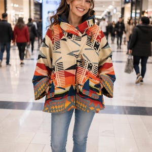 May include: A hooded jacket featuring a geometric pattern in shades of cream, orange, red, and blue. The jacket has wide sleeves, two front pockets, and is fastened with two wooden toggle closures. It is paired with light blue denim jeans and white sneakers.