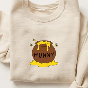 Puede incluir: Sudadera de color crema con una ilustración de dibujos animados de un tarro de miel marrón con miel amarilla goteando. La palabra "HUNNY" está impresa en el tarro. Dos pequeñas abejas vuelan cerca del tarro. La sudadera está doblada.