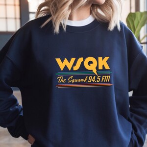 Könnte beinhalten: Marineblaues Sweatshirt mit dem Text "WSQK The Squawk 94.5 FM" in Gelb, Grün, Orange und Rot. Das Sweatshirt hat einen Rundhalsausschnitt und lange Ärmel. Das Design ist im Retro-Stil.