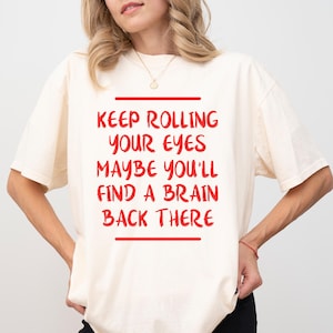 May include: Cream-colored t-shirt with the red text "KEEP ROLLING YOUR EYES MAYBE YOU'LL FIND A BRAIN BACK THERE" in a rectangular box. The shirt has short sleeves and a relaxed fit.