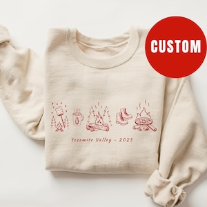 May include: Cream-colored sweatshirt with a red camping-themed graphic. The design includes a campfire, trees, boots, and a pan. The text "Yosemite Valley - 2025" is printed below the graphic. A red circle with the word "CUSTOM" is on the upper right.