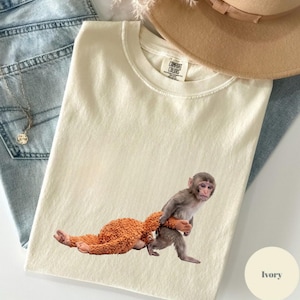 Könnte beinhalten: Elfenbeinfarbenes T-Shirt mit einem Aufdruck eines Affen, der ein Stofftier trägt. Das T-Shirt hat einen Rundhalsausschnitt und ist aus weichem Material. Das Comfort Colors-Etikett ist sichtbar. Das Shirt liegt auf einer blauen Jeans und einem beigen Hut.