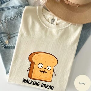 Puede incluir: Camiseta de color marfil con una ilustración de dibujos animados de una rebanada de pan con una expresión de sorpresa y las palabras "WALKING BREAD" en negro. La camiseta está doblada sobre unos vaqueros azules y un sombrero beige.