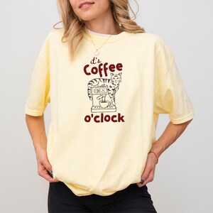 May include: A light yellow t-shirt with the phrase "it's coffee o'clock" in maroon. The graphic features a cat operating a coffee machine. The shirt is a comfortable, casual style.