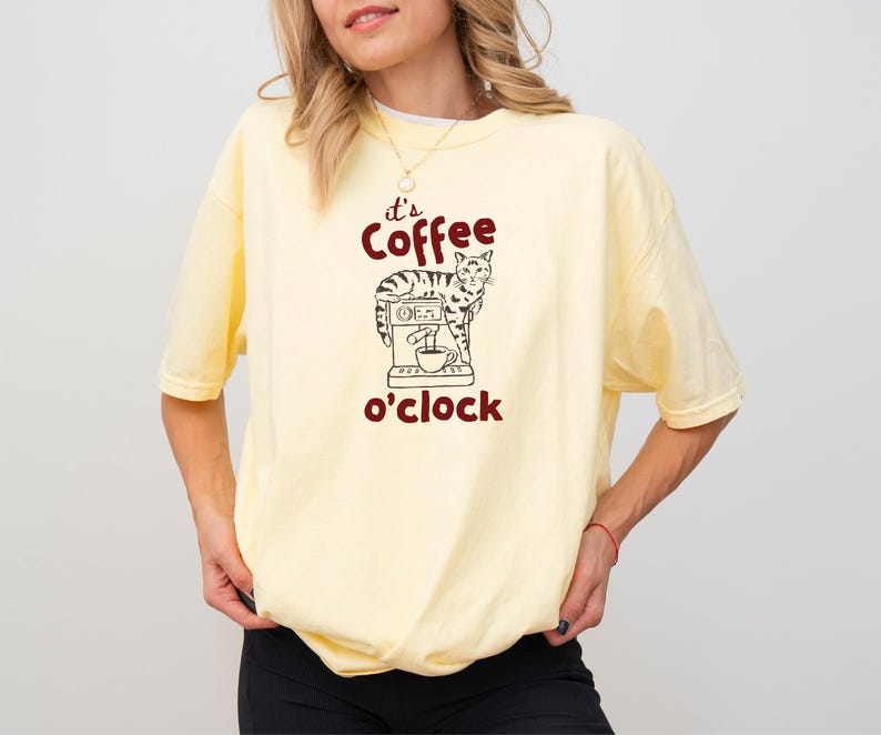 May include: A light yellow t-shirt with the phrase "it's coffee o'clock" in maroon. The graphic features a cat operating a coffee machine. The shirt is a comfortable, casual style.