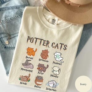 May include: Ivory-colored t-shirt with the text "POTTER CATS" and cartoon cat illustrations. The shirt features various cat characters with names like Rawrnald, Hermeownie, and Pawdamort. The shirt is made of a soft material.