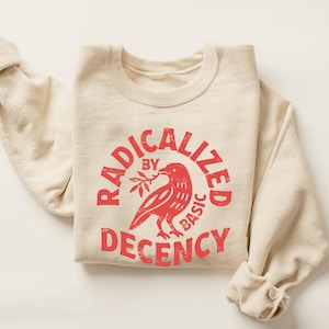 May include: Cream-colored sweatshirt with the red text "RADICALIZED BY BASIC DECENCY" in a circular design. A red bird holding a sprig is in the center. The sleeves are cuffed.