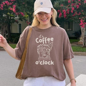 May include: Brown t-shirt with the text "It's Coffee o'clock" and a graphic of a cat and coffee machine. The shirt is worn with a beige cap, gold hoop earrings, and a brown shoulder bag. The person is smiling.